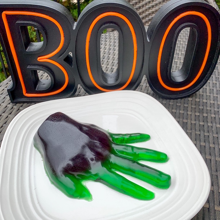 Giant gummy recipe for Halloween finished product of a green and purple gummy hand on a plate with a sign that says boo